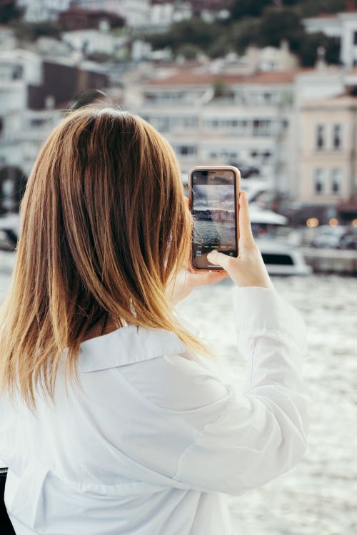 Free Woman capturing waterfront scenery with smartphone. Urban harbor backdrop. Stock Photo