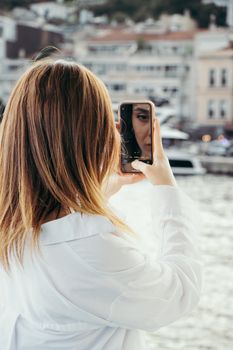 A woman captures a selfie with her phone by a city waterfront, featuring a scenic urban backdrop.