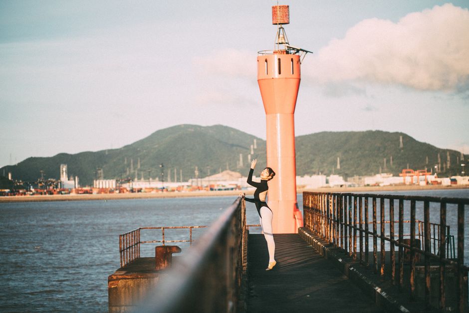 A ballet dancer poses artistically on a pier at sunrise, with scenic coastline and lighthouse in view.