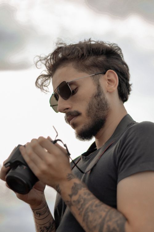 Free A bearded man with sunglasses and tattoos examines his camera outdoors. Stock Photo