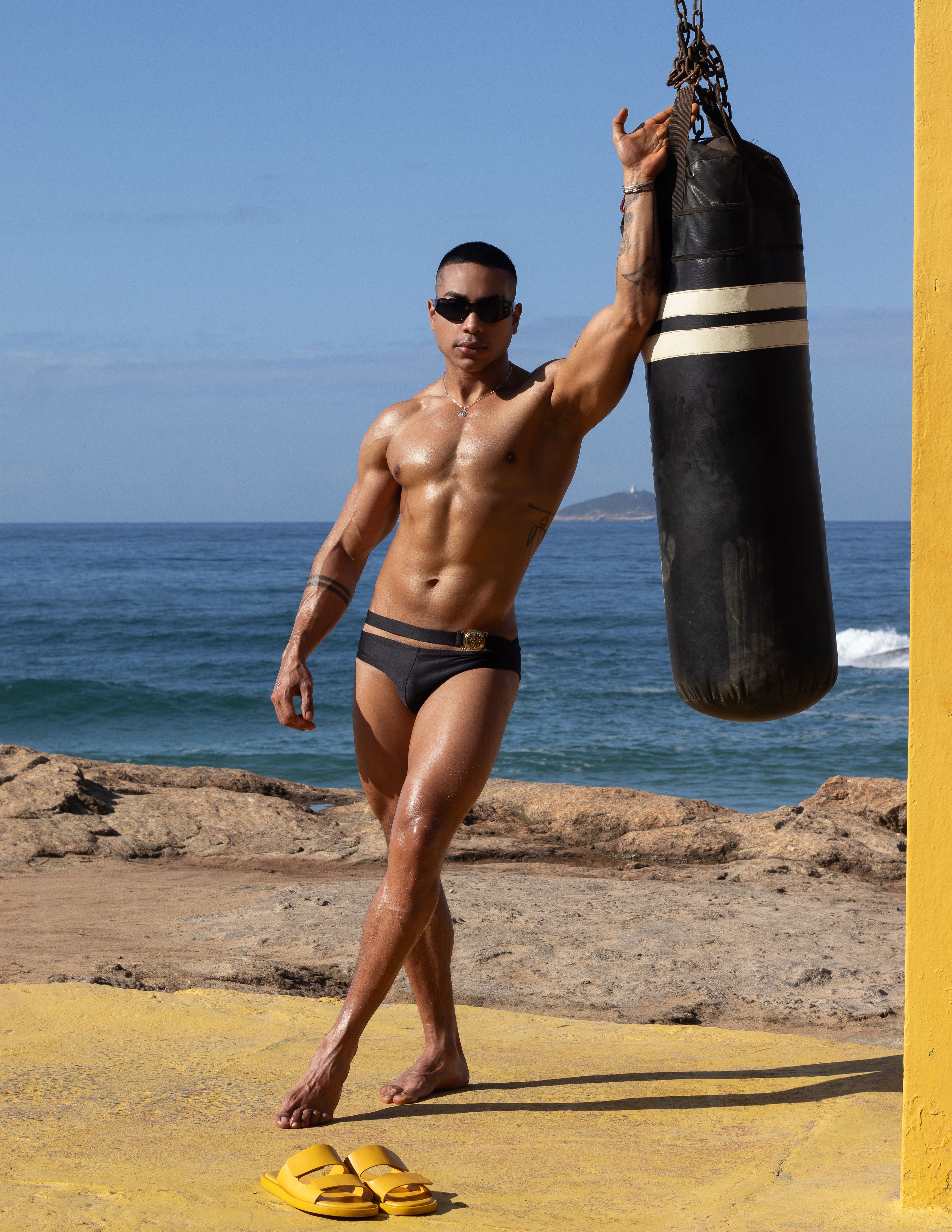 Homem musculoso de maiô posa com saco de pancadas em uma ensolarada praia do Rio de Janeiro.