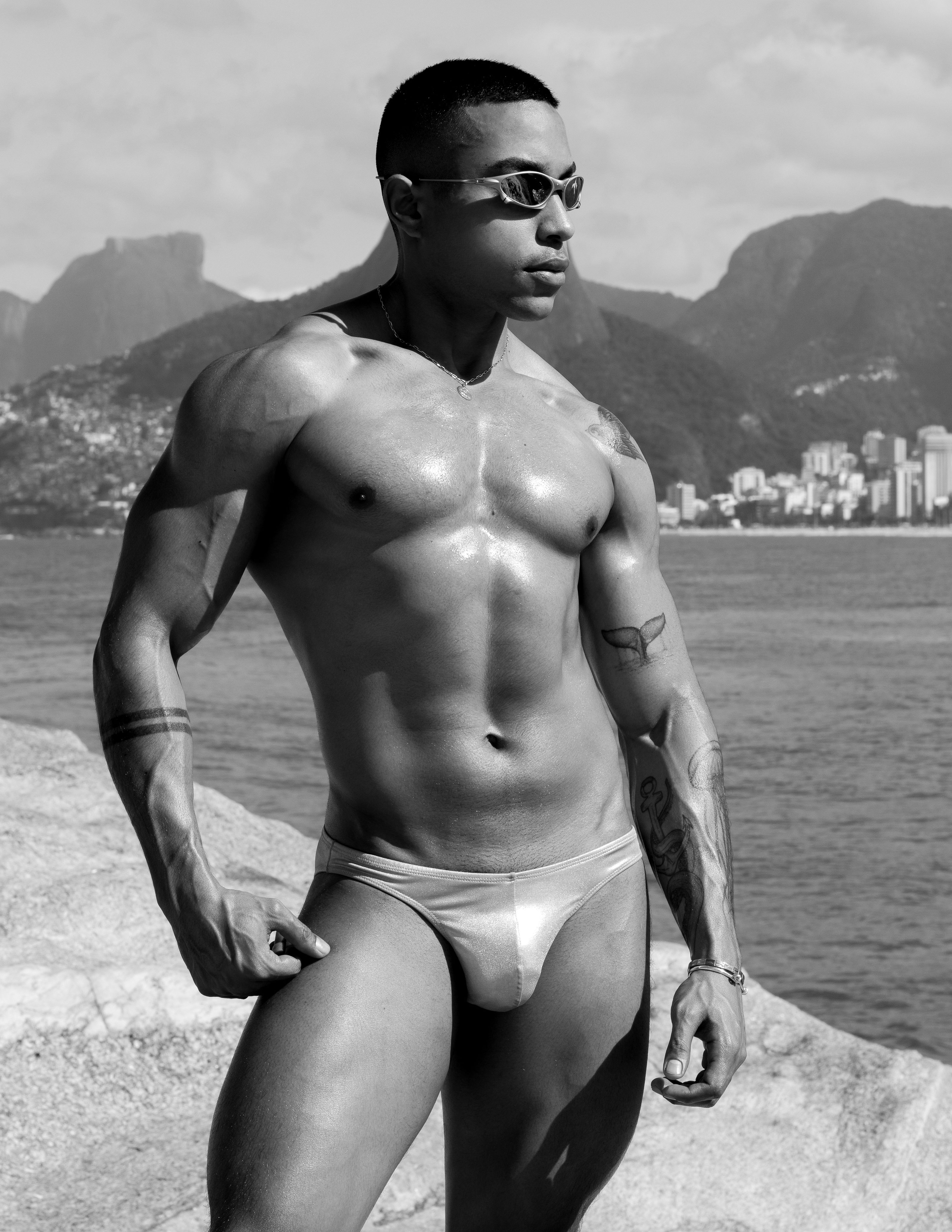Retrato em preto e branco de um homem musculoso em pé na orla do Rio de Janeiro, exibindo tatuagens e força.