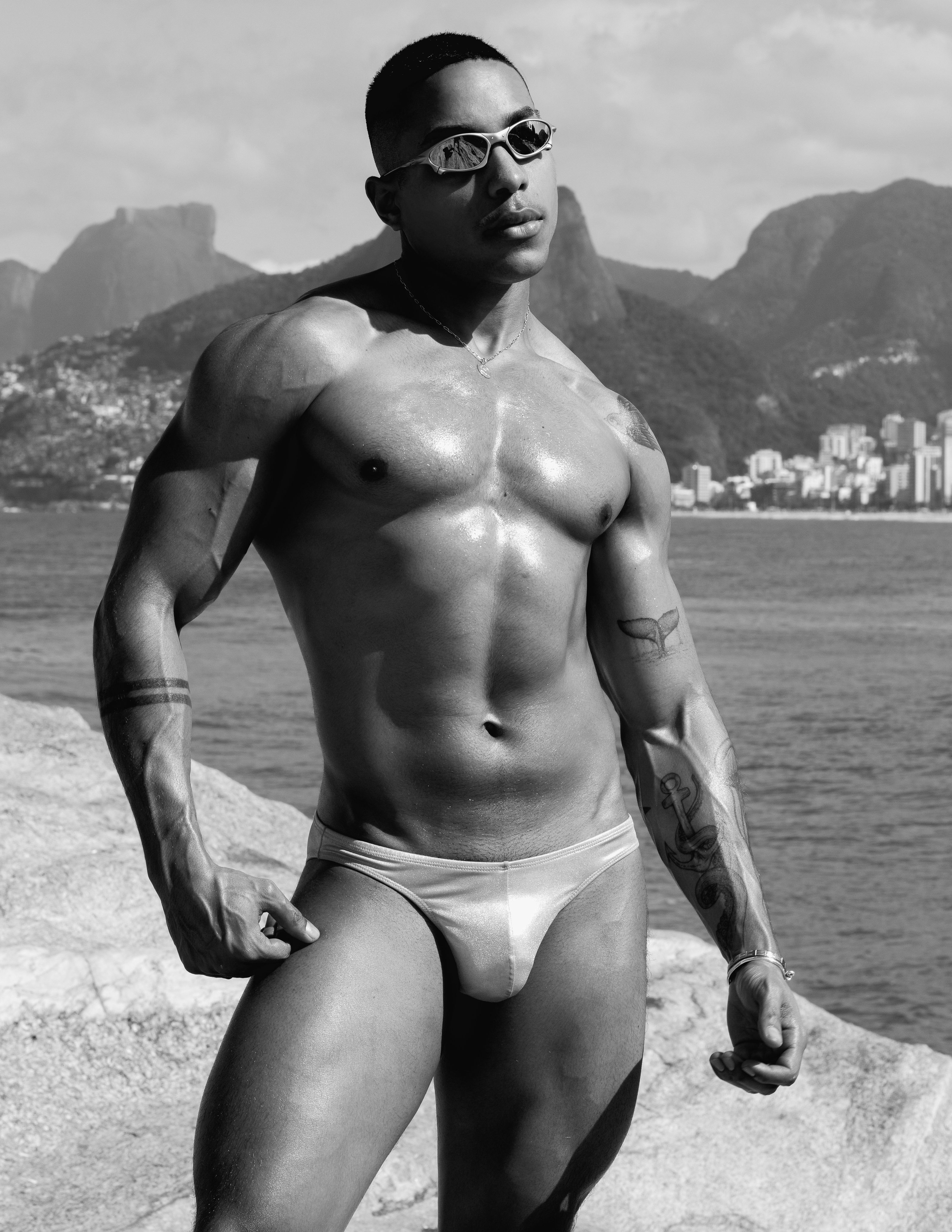 Um homem musculoso em sunga posa confiantemente na costa do Rio de Janeiro. Preto e branco.