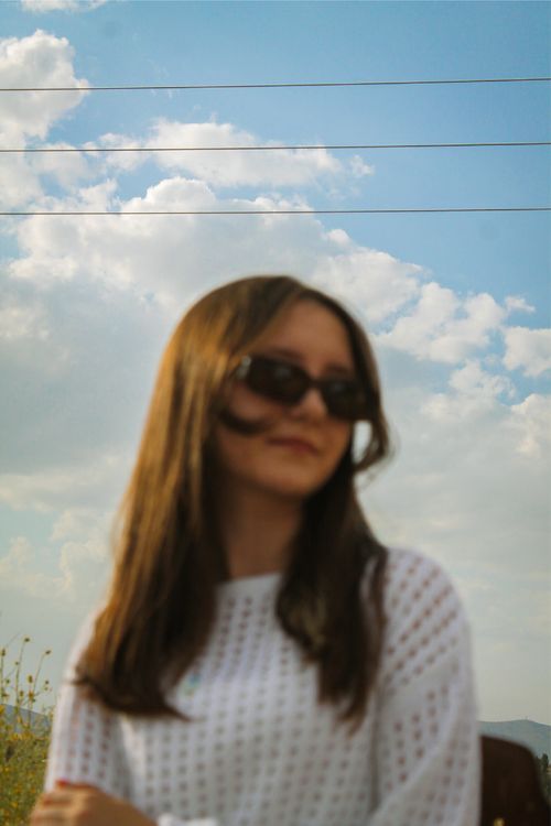 Free A woman with sunglasses is softly blurred against a vibrant blue sky with clouds. Stock Photo
