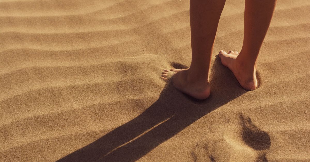 Close-up of a person standing barefoot on sunlit sand with visible footprints.