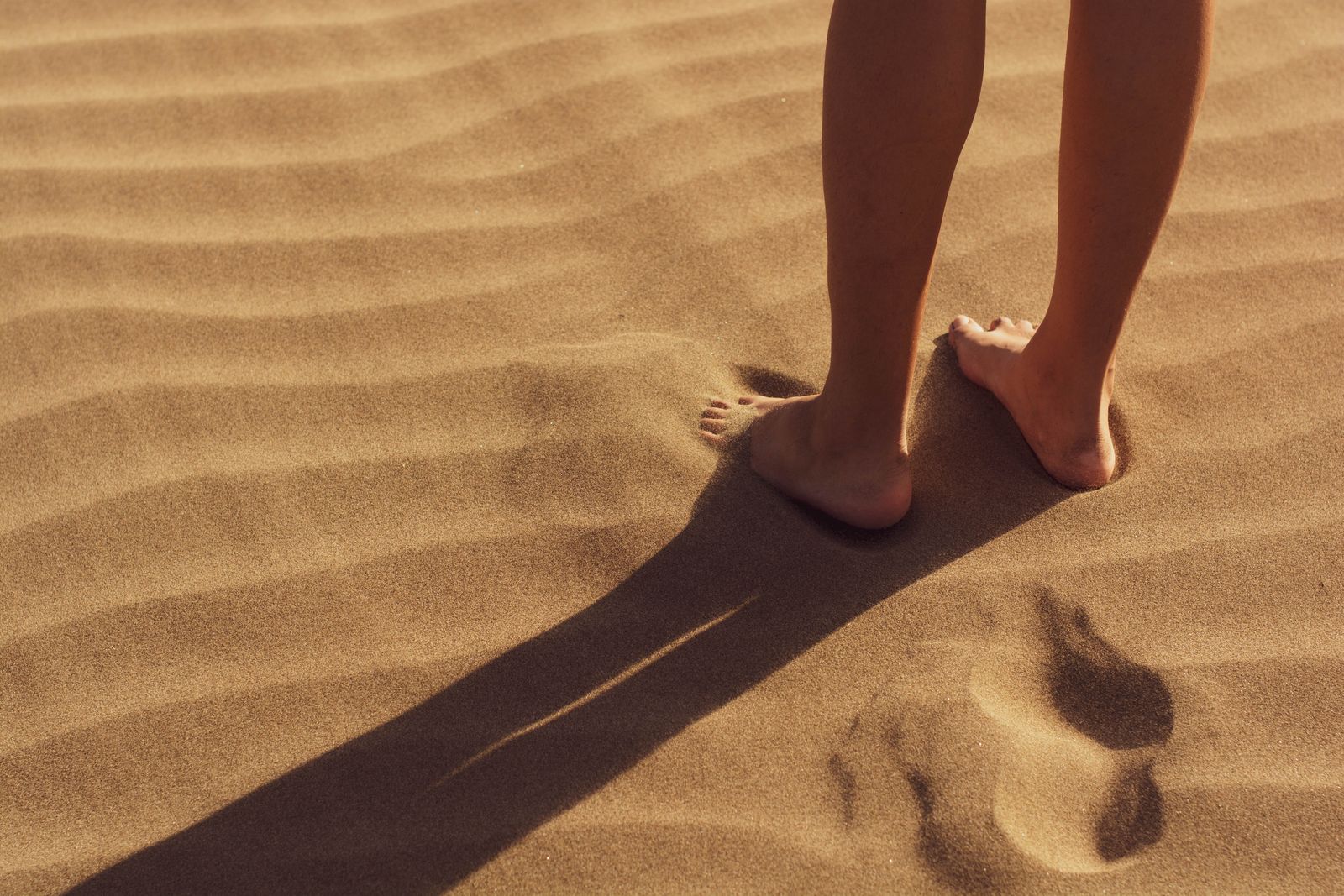 Bare feet and legs standing on rippled golden sand, casting a long shadow in warm sunlight.