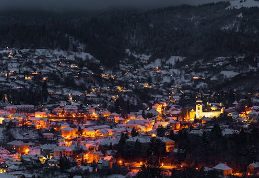 A scenic aerial view of a snowy town with warm lights glowing at night, surrounded by forested hills.