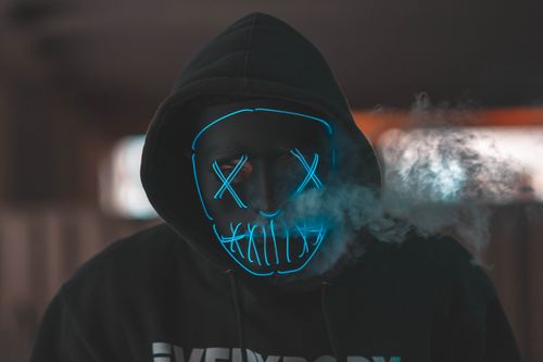 Free Hooded figure with a neon mask emitting smoke, creating a mysterious atmosphere. Stock Photo