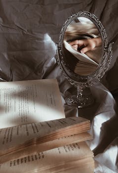 A vintage mirror reflecting hands holding an open book, surrounded by pages.