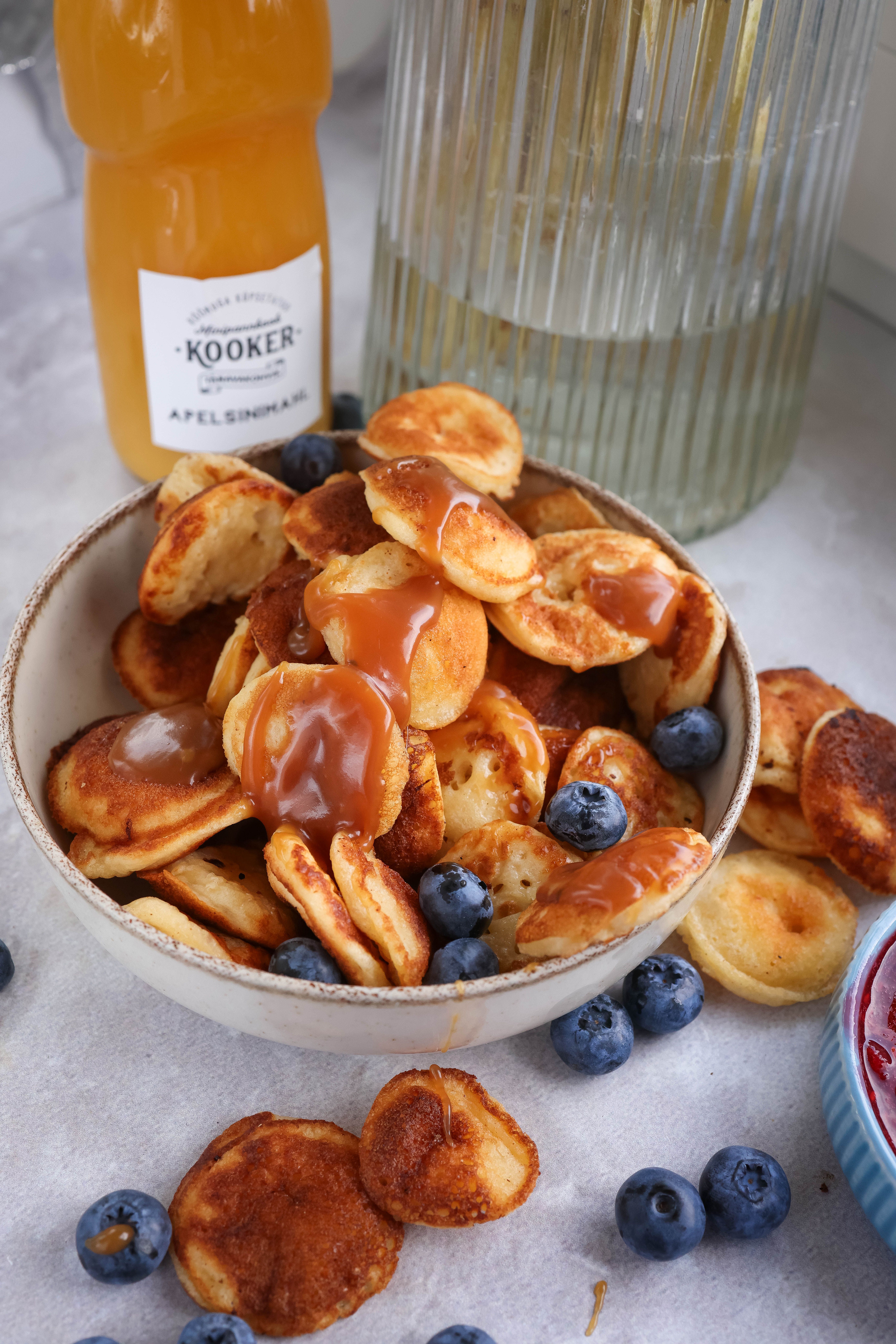 A small stack of golden mini pancakes about two inches wide, drizzled with maple syrup and topped with fresh blueberries on a white plate.