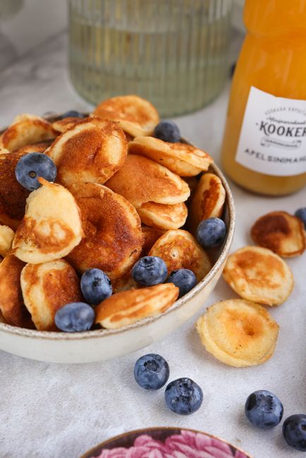 A dimpled cast iron poffertjes pan filled with small golden-brown mini pancakes, one plate dusted heavily with powdered sugar and topped with a pat of melting butter