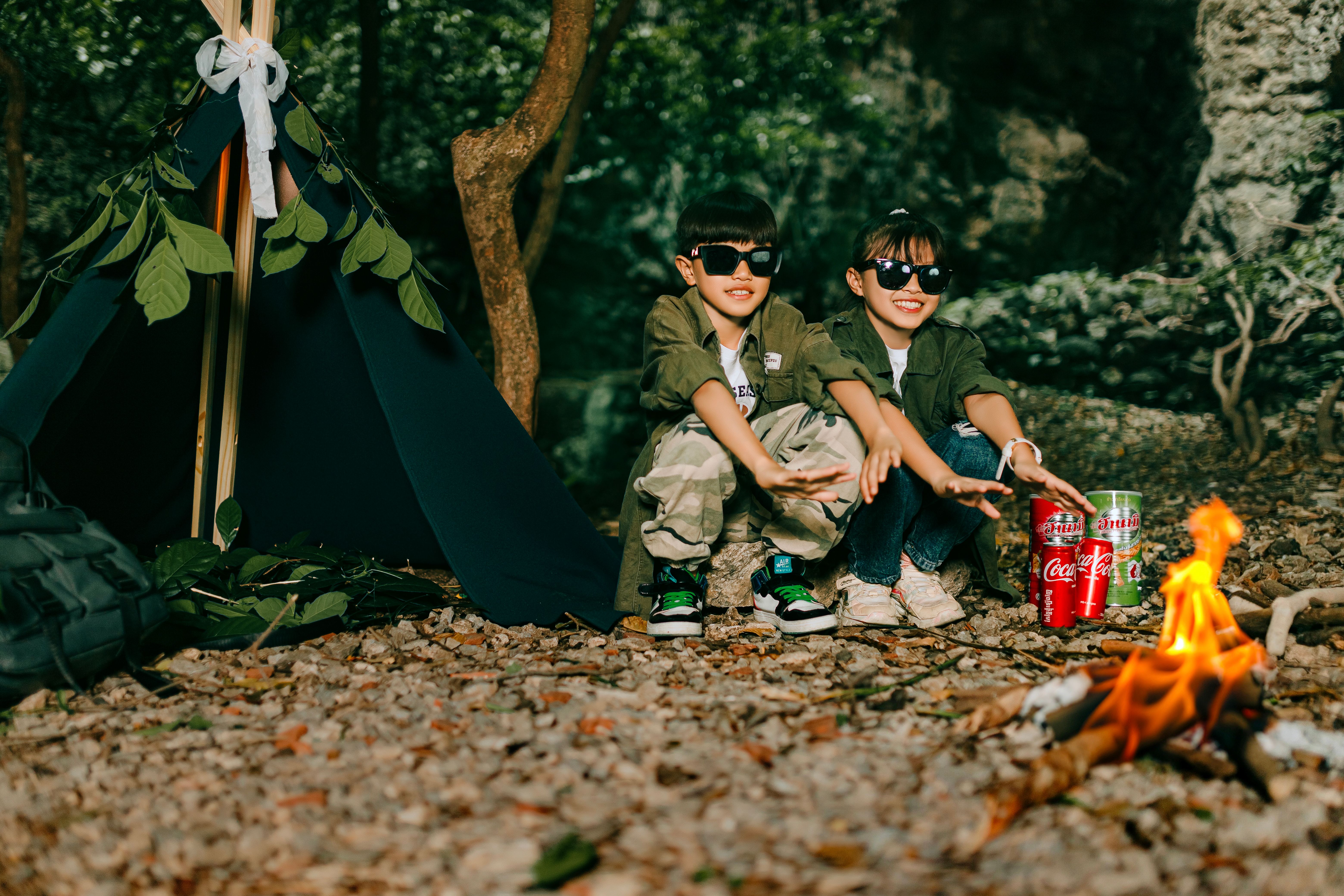 Two cheerful children enjoy a campfire adventure outdoors, sitting by a tent with drinks.