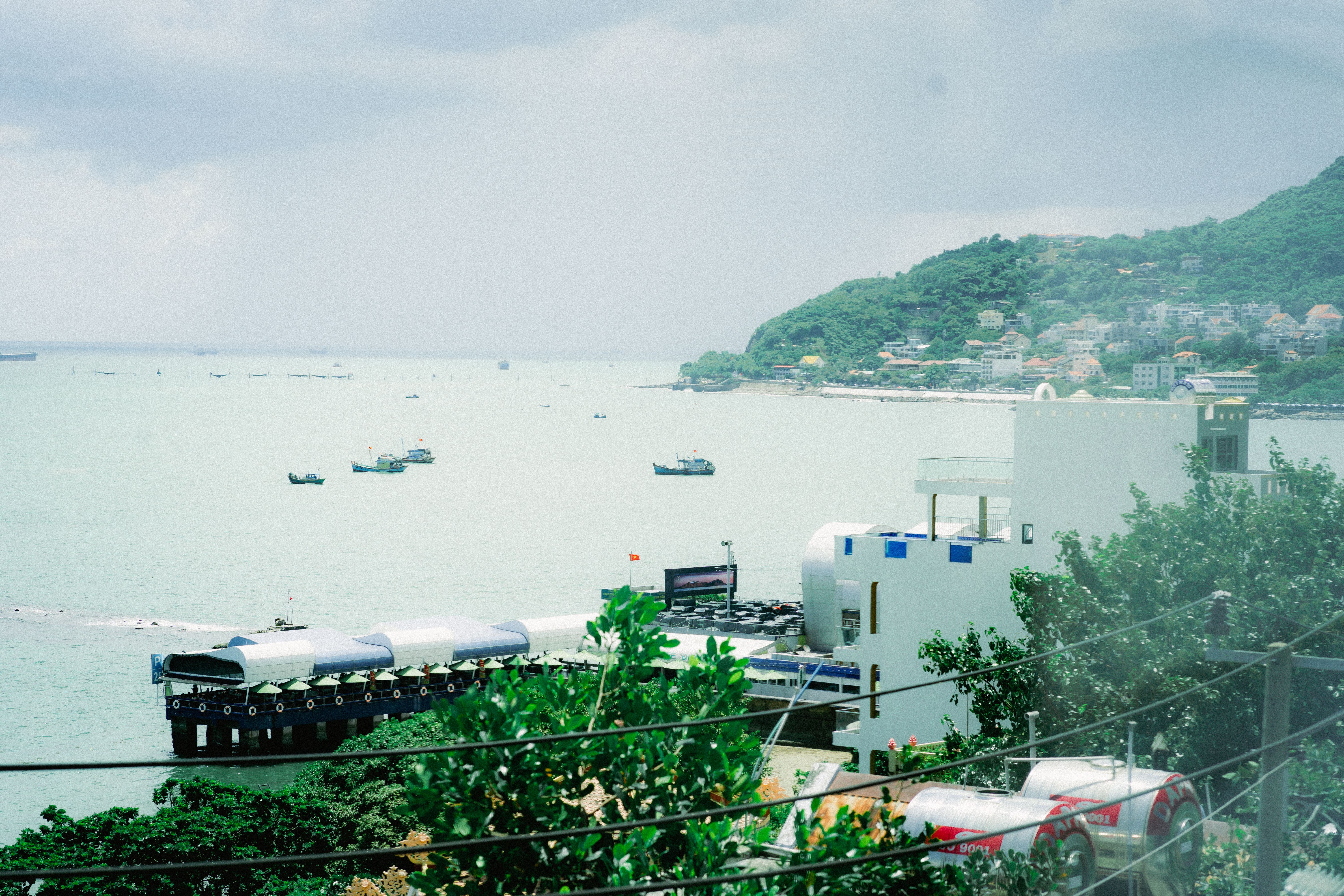 A scenic aerial view of a coastal town with buildings, sea, and green hillside in summer.