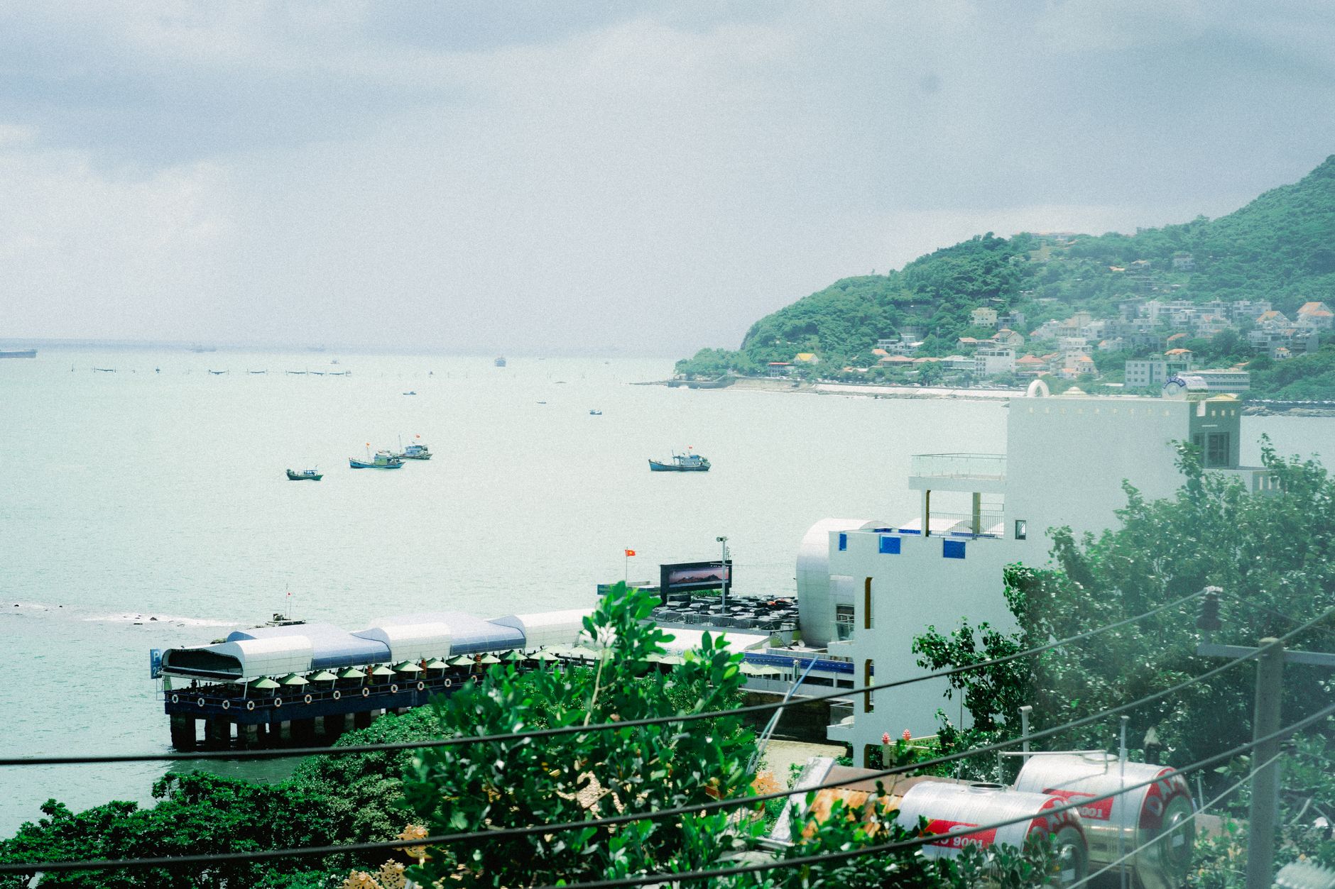 A scenic aerial view of a coastal town with buildings, sea, and green hillside in summer.