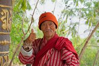 A senior Bhutanese man wearing traditional red clothing and an orange hat gives a thumbs up outdoors.