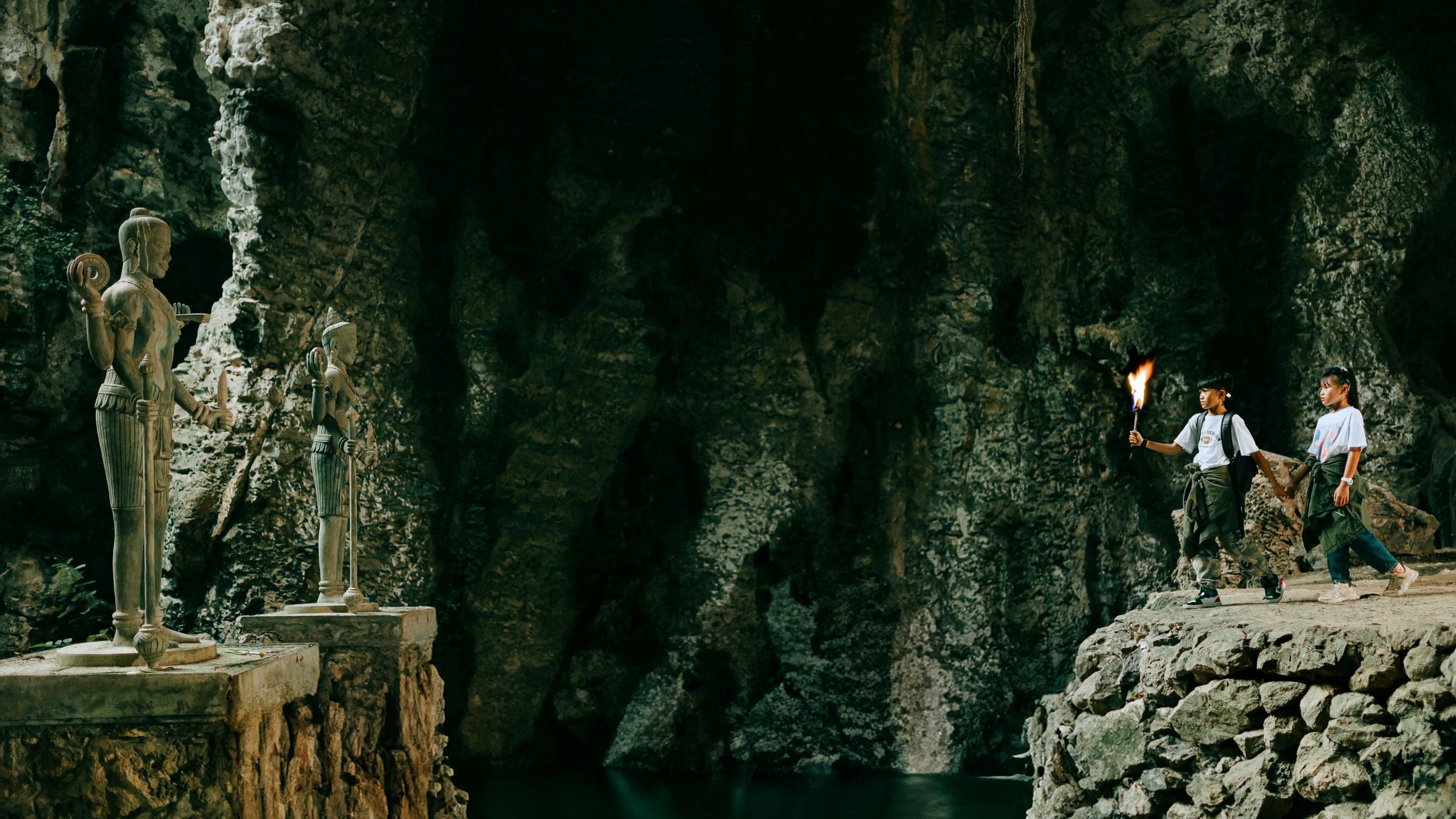 Two teenagers explore a cave with historical statues, lighting the way with a torch.