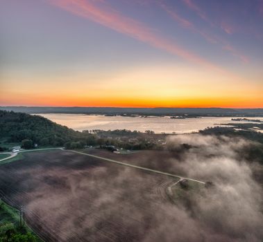A stunning aerial shot capturing fog over rural farmland with a colorful sunrise over a distant lake.