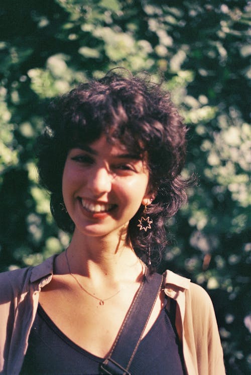 Photo of a Young Smiling Woman Standing on the Background of Green Trees 