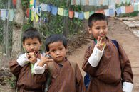 Three Bhutanese boys in traditional attire posing with peace gestures outdoors on a path with prayer flags.