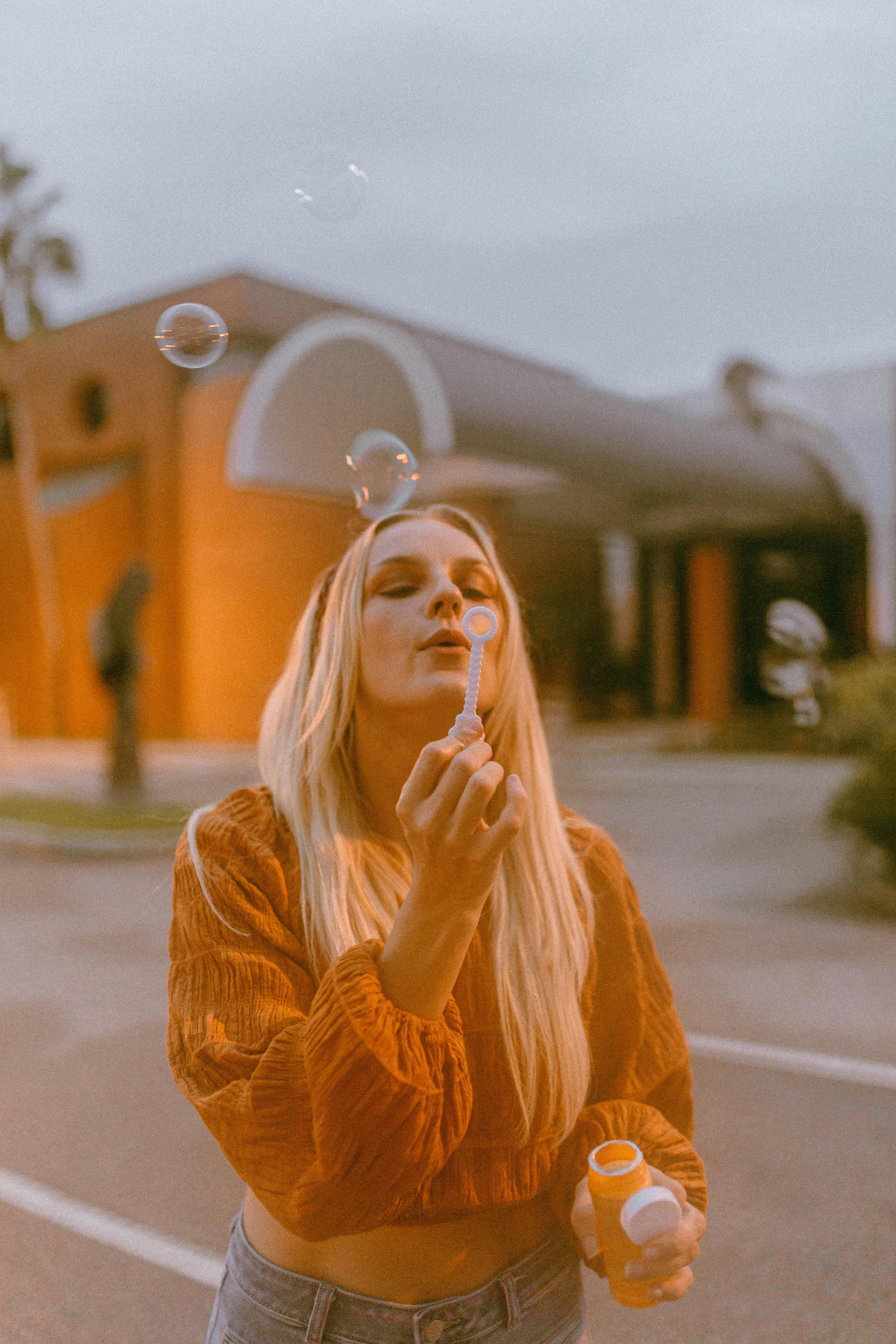 Blonde woman with long hair blowing bubbles on city streets, urban fashion style.
