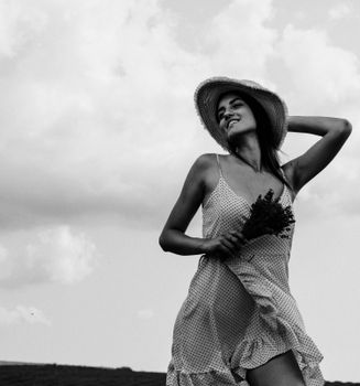 Cheerful woman in a hat holding flowers in a rural setting, black and white.