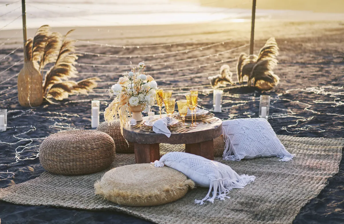 A styled luxury beach picnic at golden hour with cushions, florals, and glassware on the sand