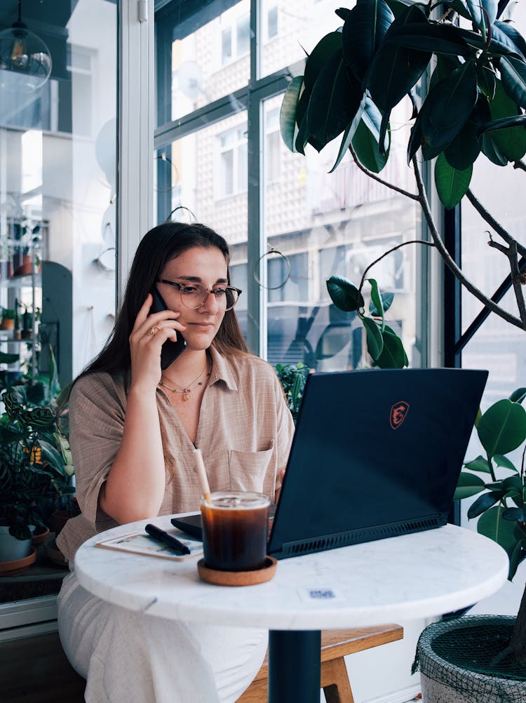 Young Woman Sitting At The Table In A Cafe And Using A Laptop And A Smartphone 