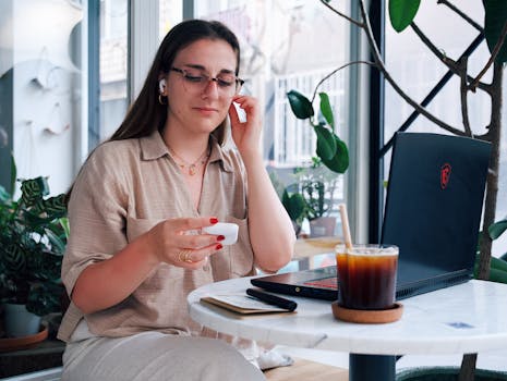 Woman sitting in a cafe with iced coffee, using earbuds next to a laptop.