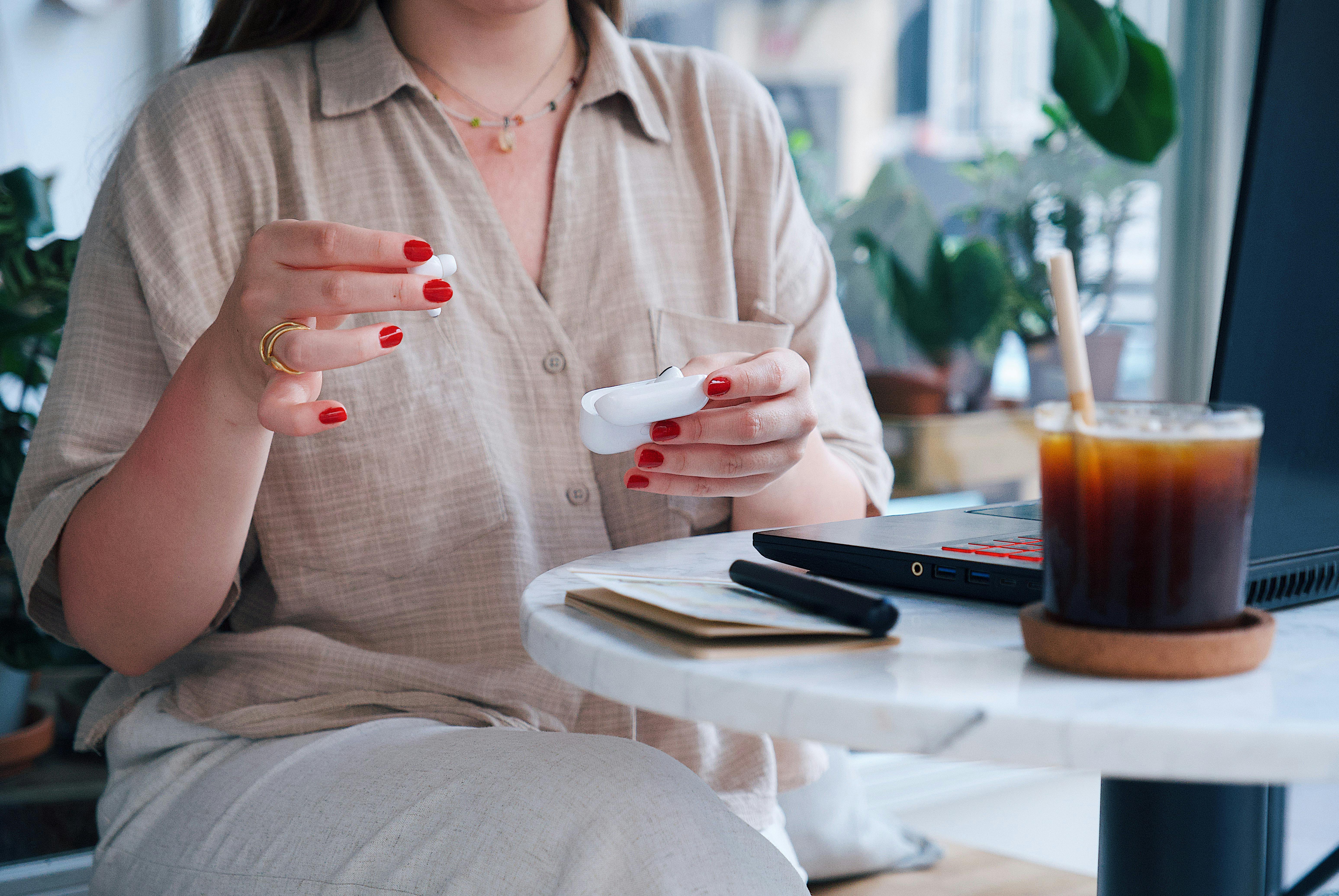 Woman handling wireless earbuds at a cafe table with iced coffee and a laptop, enjoying leisure time.