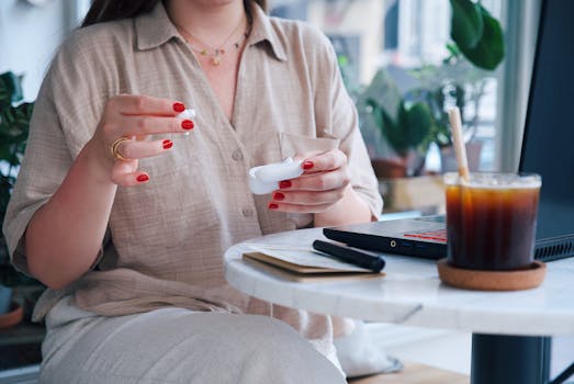 Woman handling wireless earbuds at a cafe table with iced coffee and a laptop, enjoying leisure time.