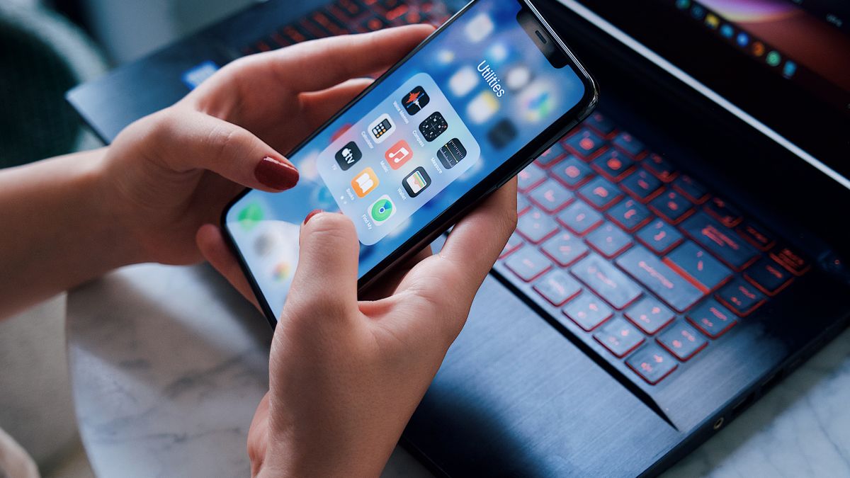 Close-up of a woman holding a smartphone showing utilities apps while sitting by a laptop indoors.