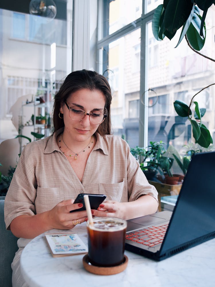 Young Woman Sitting At The Table In A Cafe And Using A Laptop And A Smartphone 