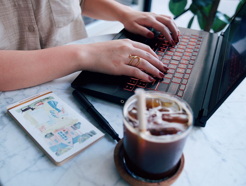 Jasper AI vs Copy AI: Bulk Social Caption Creation Myths Debunked A woman typing on a laptop at a marble table with an iced coffee and a notepad.