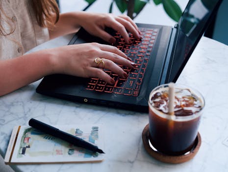 Close-up of a woman typing on a laptop with an iced coffee on the table, suggesting a casual work environment.