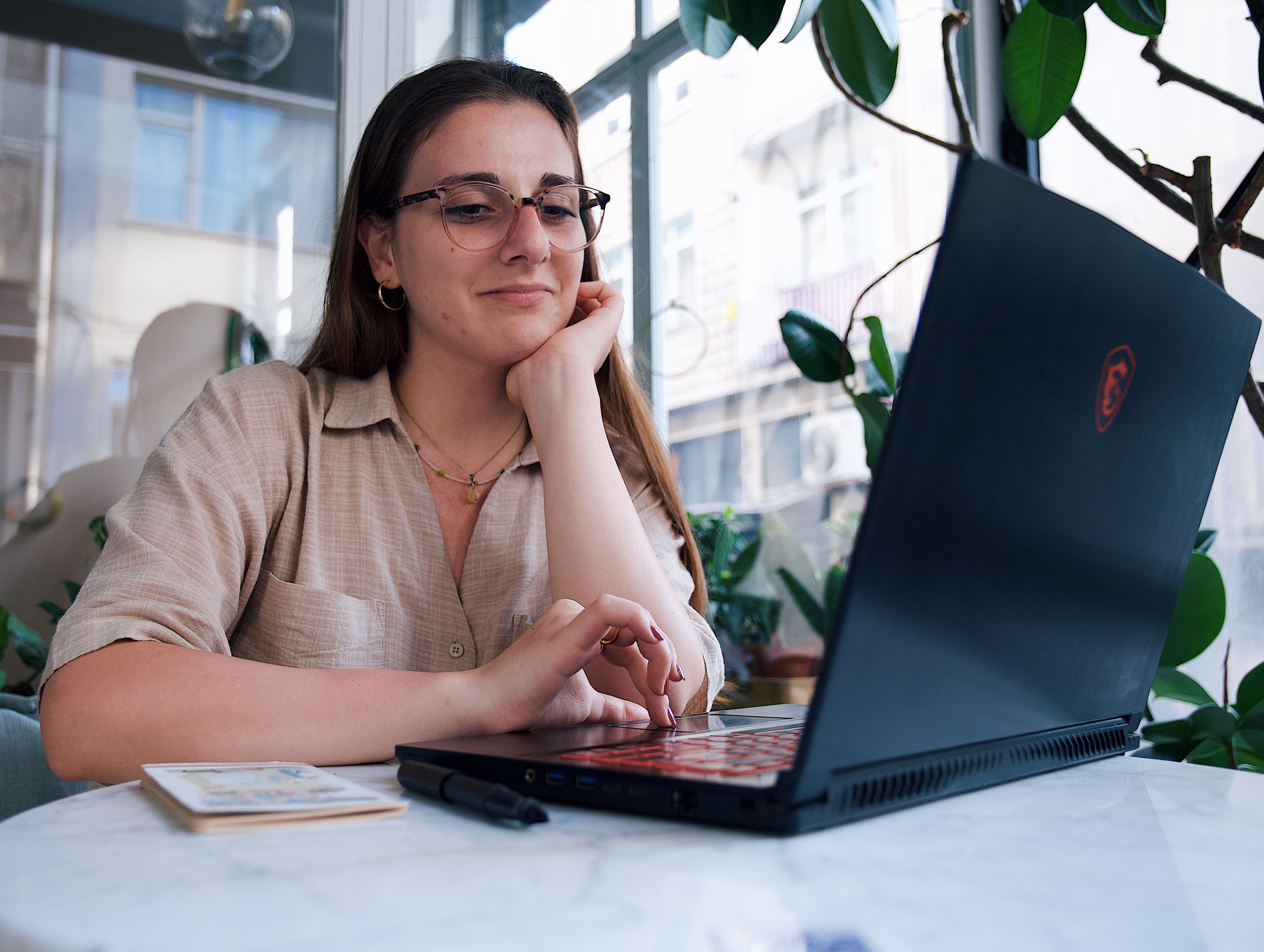 A young woman enjoys her time at a café working on her laptop, surrounded by greenery and natural light.