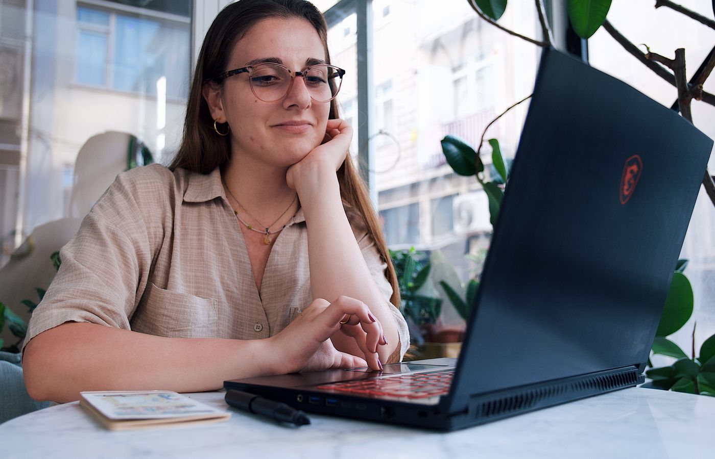 A young woman enjoys her time at a café working on her laptop, surrounded by greenery and natural light.
