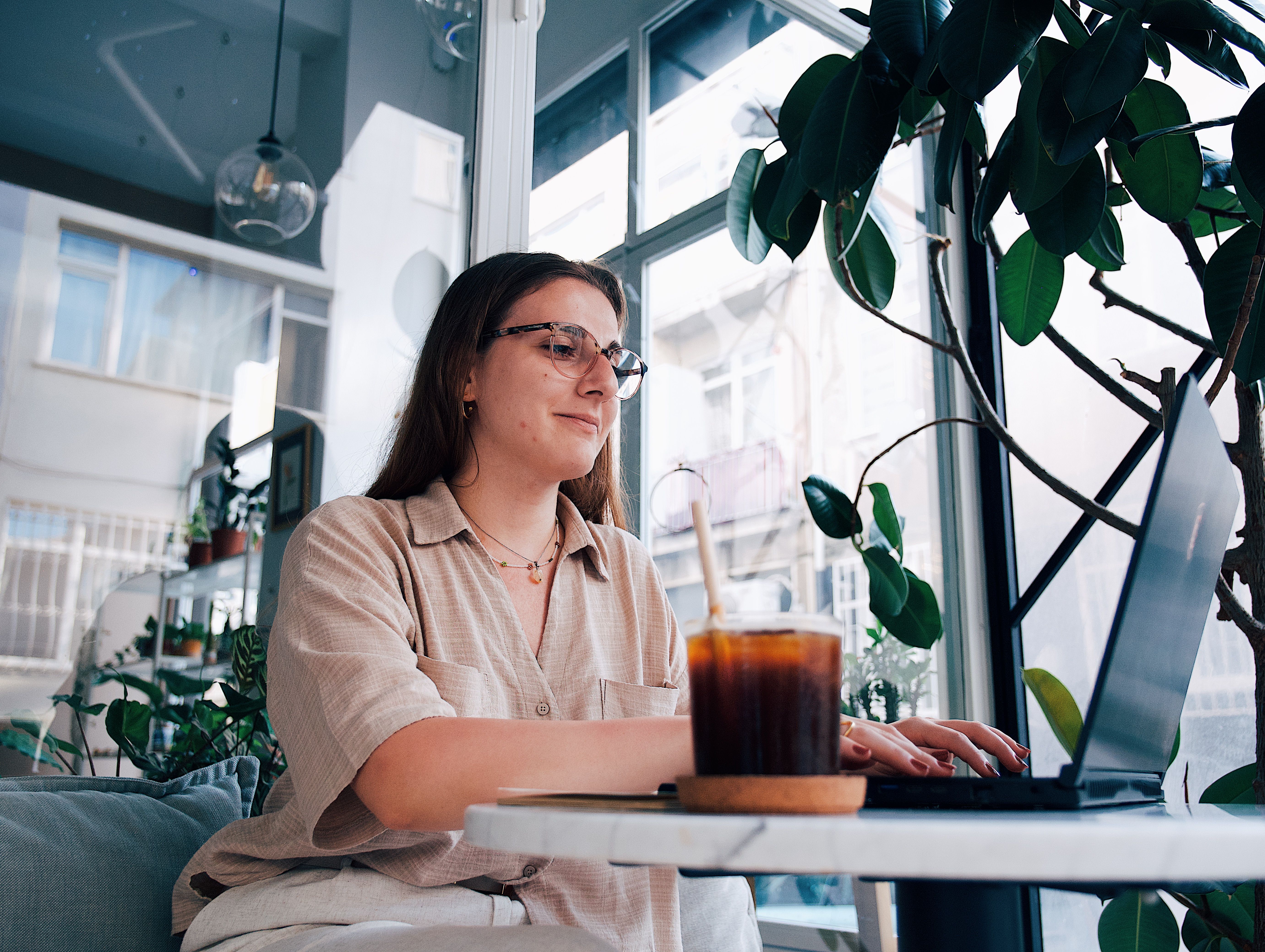 A young woman using a laptop in a stylish, sunny café with greenery and a drink.