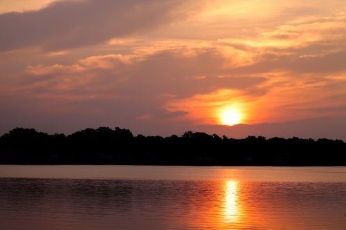 Free Beautiful sunset over a calm lake with forest silhouette, reflecting orange hues across the water. Stock Photo