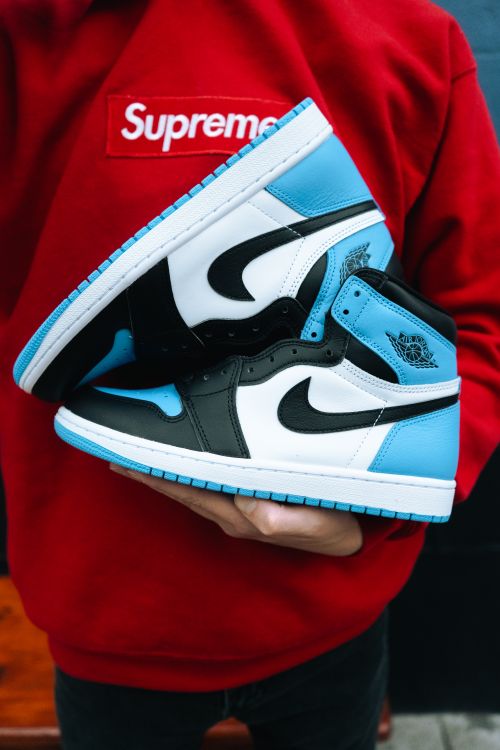 Free A person holds a stylish pair of Air Jordan sneakers against a red Supreme hoodie. Stock Photo