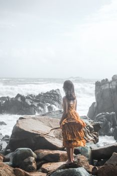 A woman in a yellow dress stands on a rocky coast, gazing at the ocean waves under a cloudy sky.