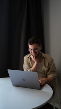 A young man smiling while using a laptop at a table indoors.