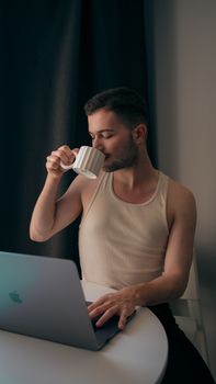 A young man in a tank top enjoys coffee while working on a laptop indoors.