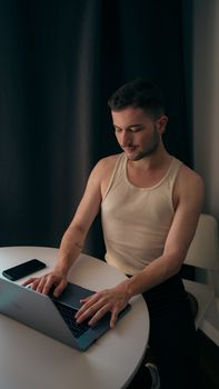 Adult man in tank top using laptop at a table, suggesting casual work or study setting indoors.