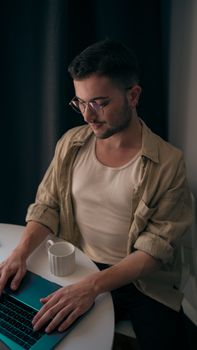 Young man with eyeglasses working on a laptop, sitting at a round table indoors with a mug.