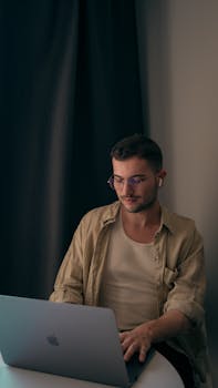 A young adult man wearing eyeglasses works on a laptop indoors, focused and engaged.