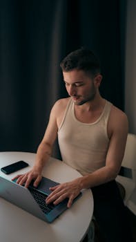 Young man in a tank top working on a laptop at a round table with a phone nearby.