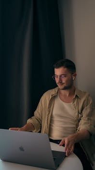 Young Caucasian man with eyeglasses working on a laptop at a table indoors.