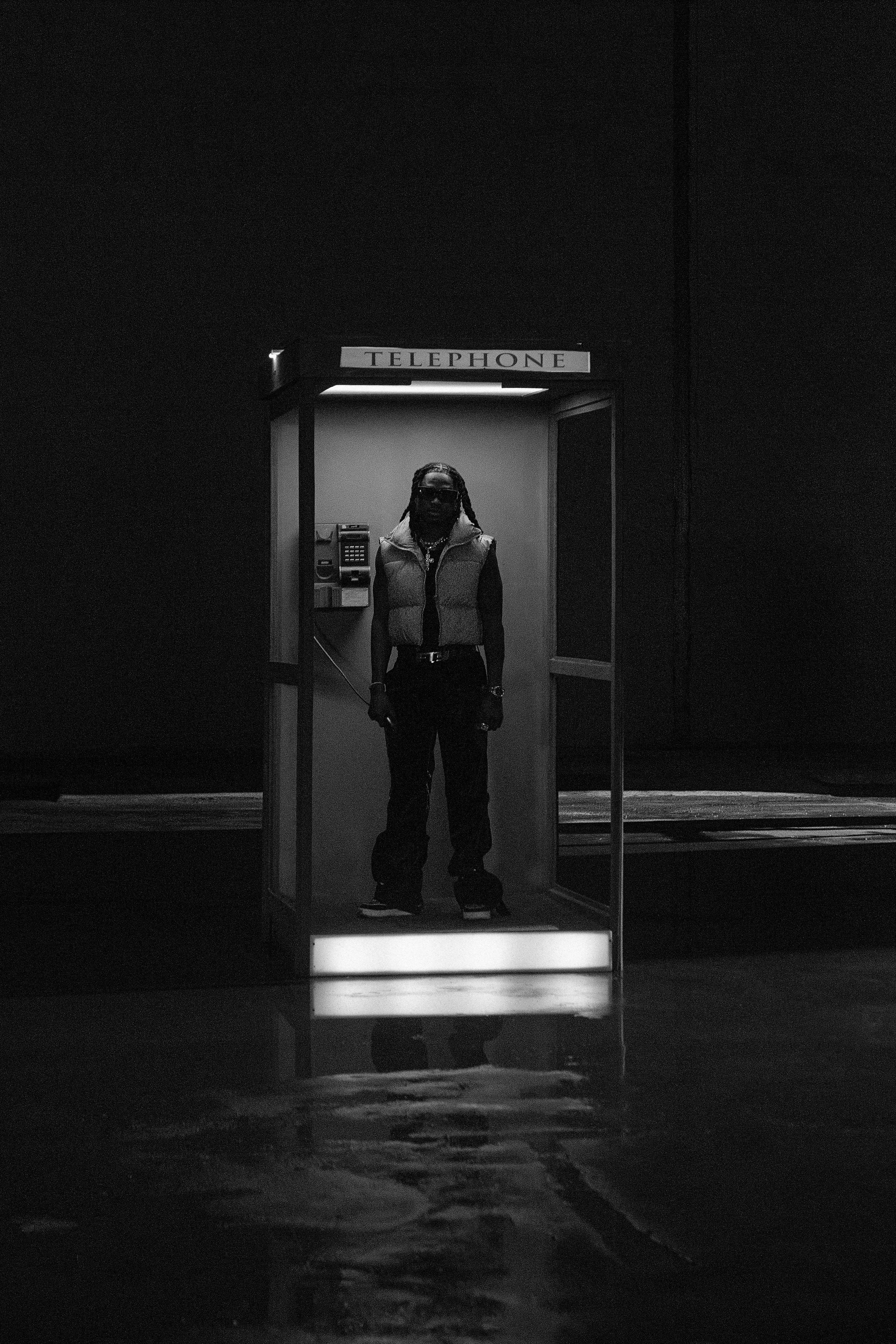 Black and white image of a person standing in a lit phone booth at night.