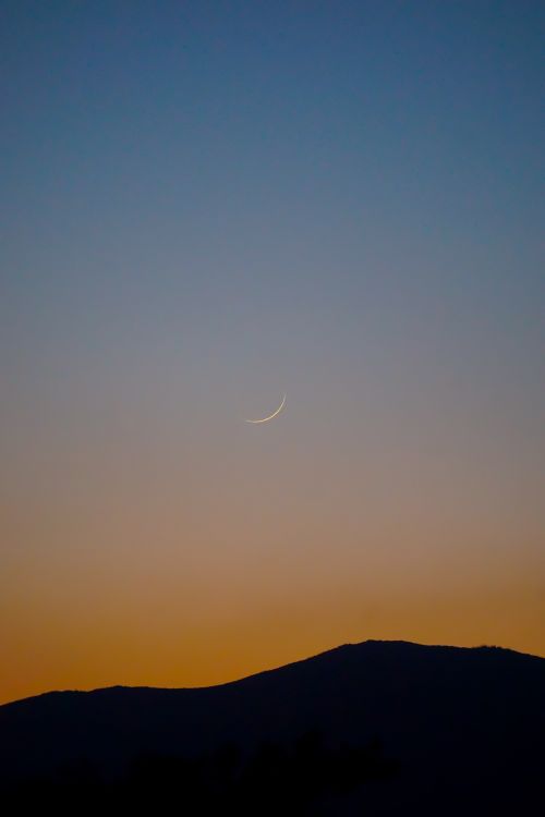 Free A serene crescent moon in a clear sky above a silhouetted hill during a vibrant sunset. Stock Photo