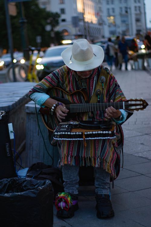Free A street musician in a hat and poncho playing guitar on a busy urban sidewalk. Stock Photo